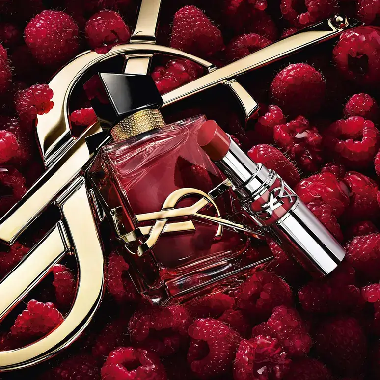 yves saint laurent libre berry crush raspberries شیشهی لیبر بری کراش در میان تمشکهای تازه و نشان طلایی YSL، کنار یک رژ لب ایو سن لوران